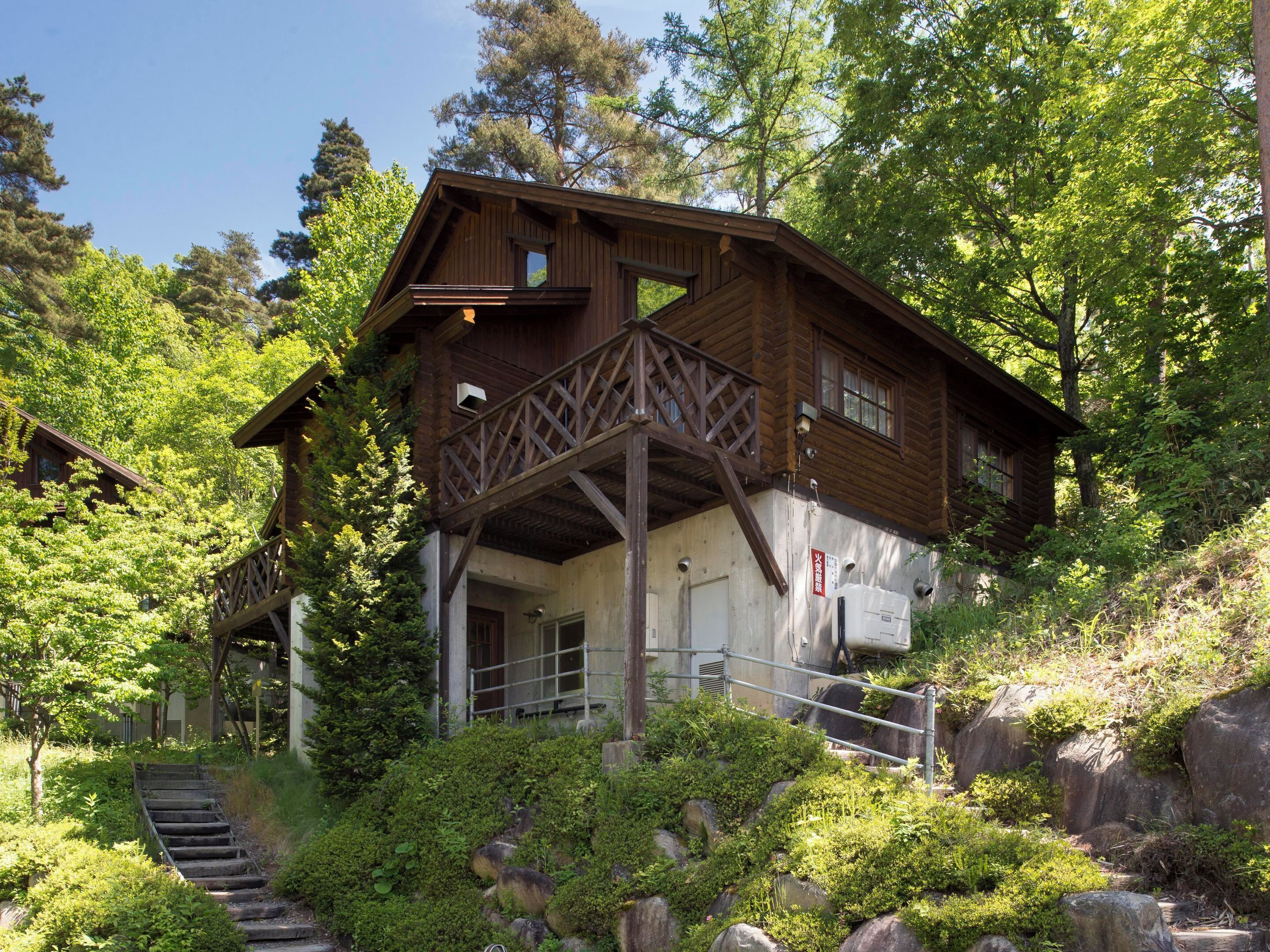 A wooden cabin with a balcony, surrounded by lush greenery and a stone staircase.