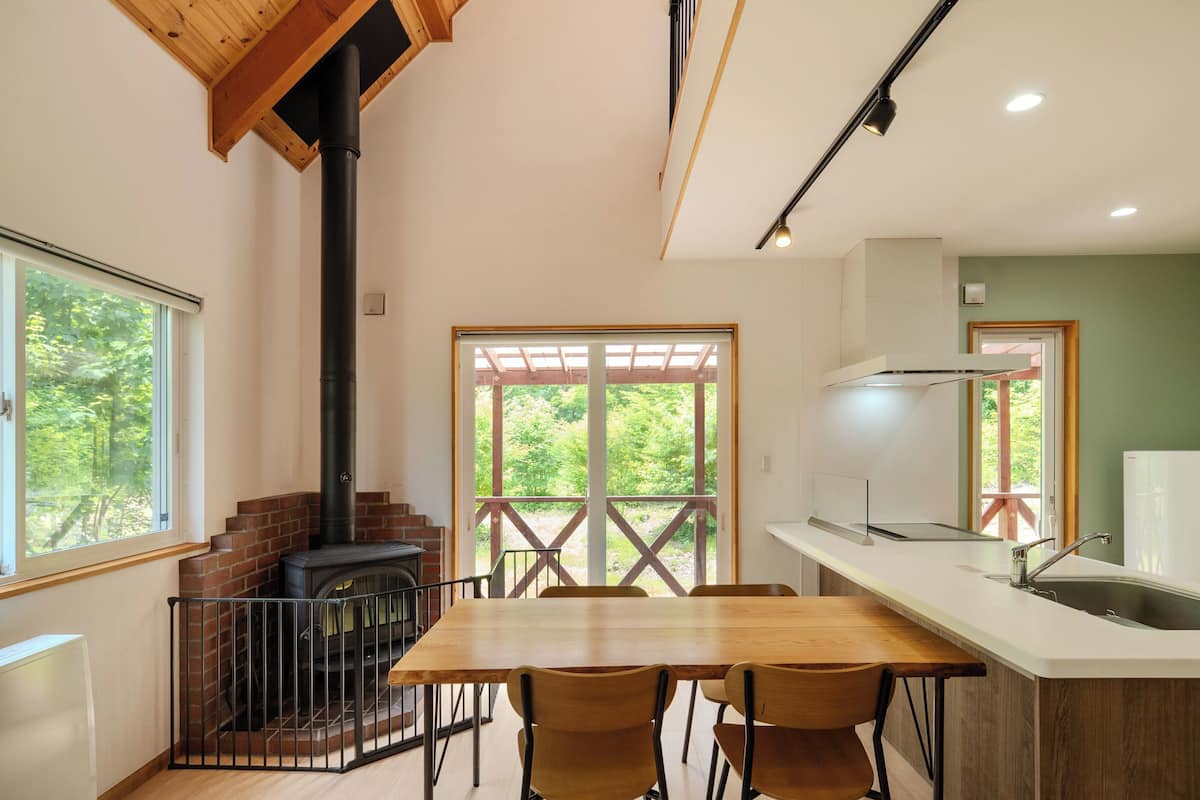 A modern kitchen with a central island, a dining area with chairs, a built-in stove, and large windows offering a view of greenery.