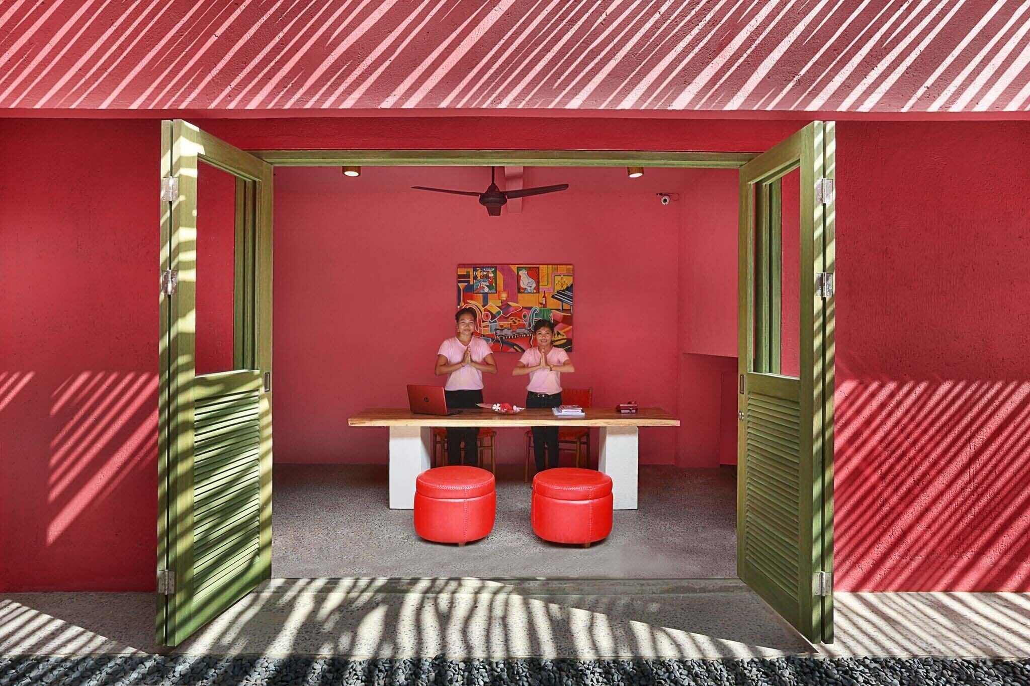 A room with a red wall, a ceiling fan, a table with two red ottomans, and a colorful painting on the wall.