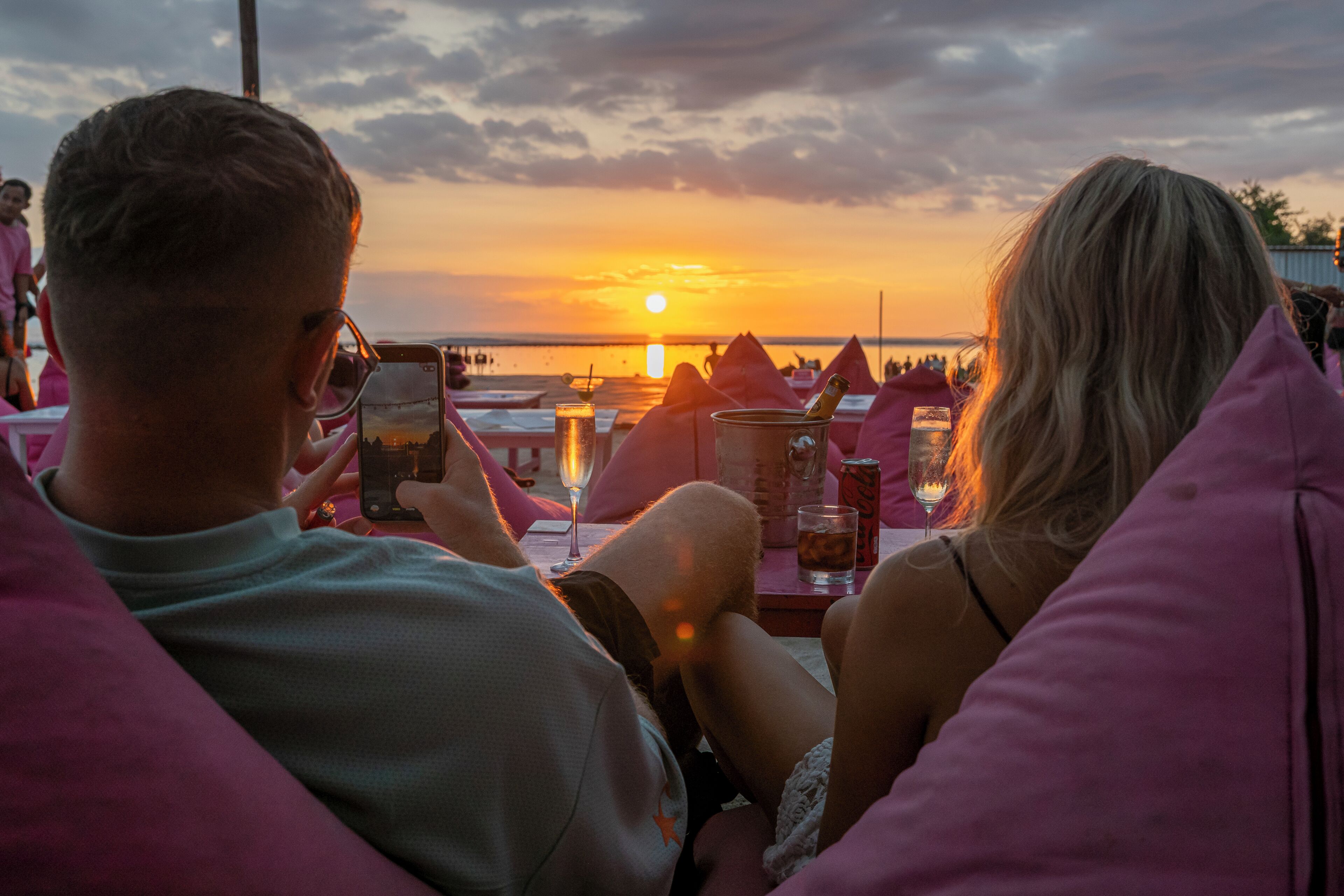 A couple seated at a table with a sunset view, a drink, and a bottle.