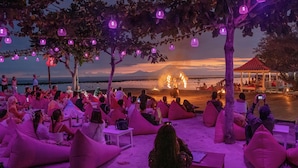 Une rencontre conviviale sur la plage, avec des personnes assises sur des poufs, un spectacle de jonglerie avec le feu et une vue sur l’océan et les montagnes.