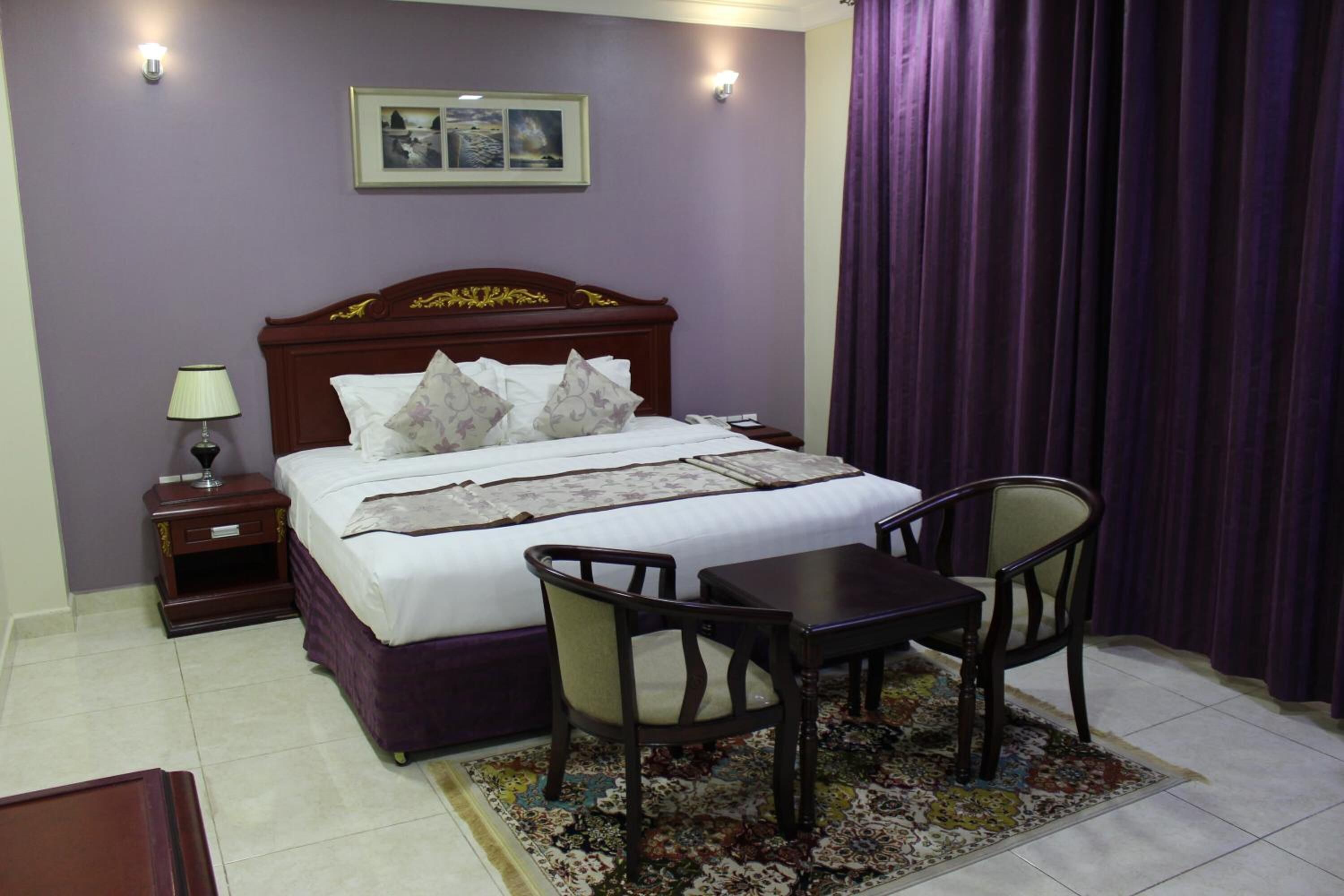 Quarto bem arrumado com cama, mesa de cabeceira, abajur, cadeira e uma mesinha.