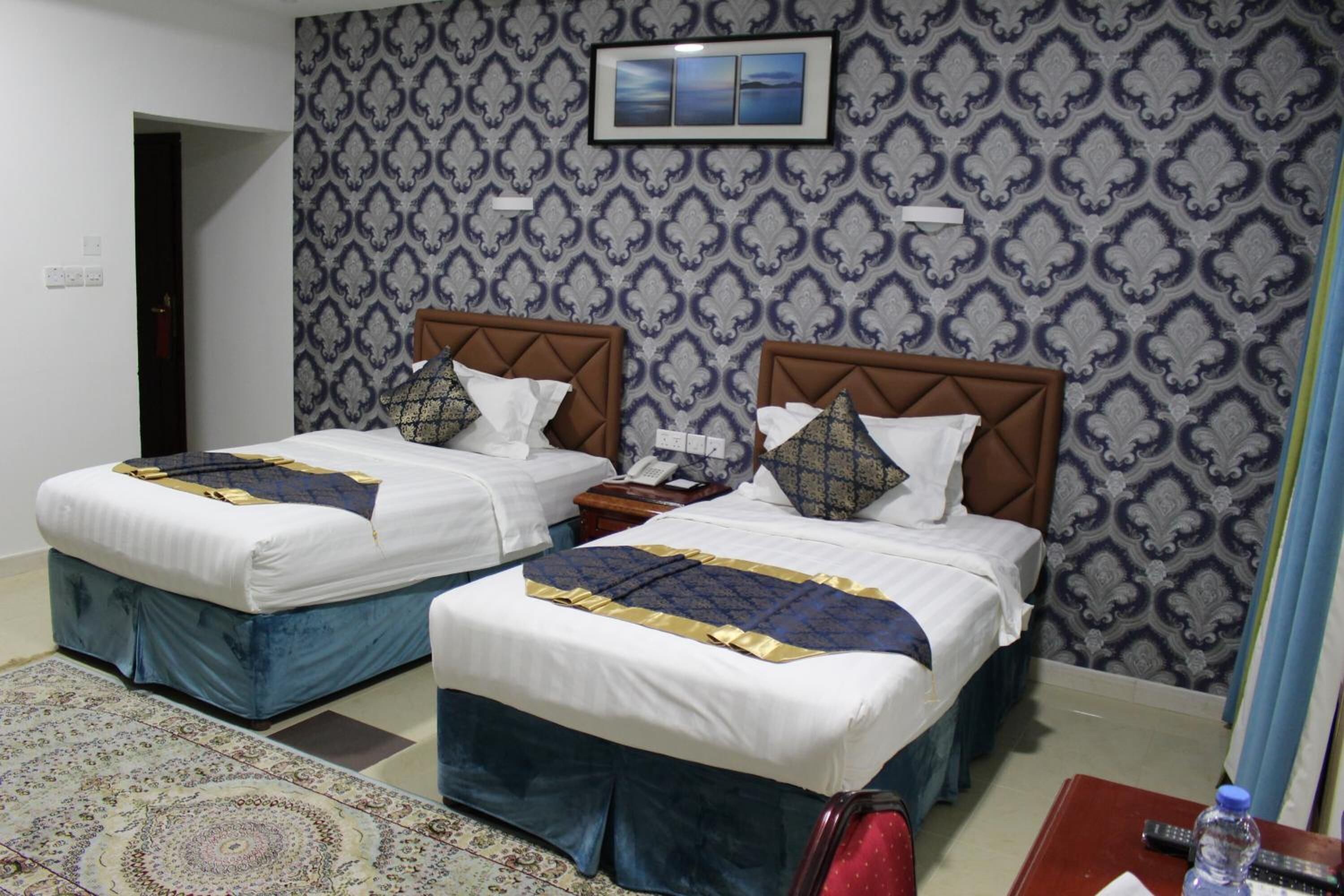 Habitación de hotel con dos camas, un papel tapiz estampado, un televisor pequeño en la pared y una maleta roja en el suelo.