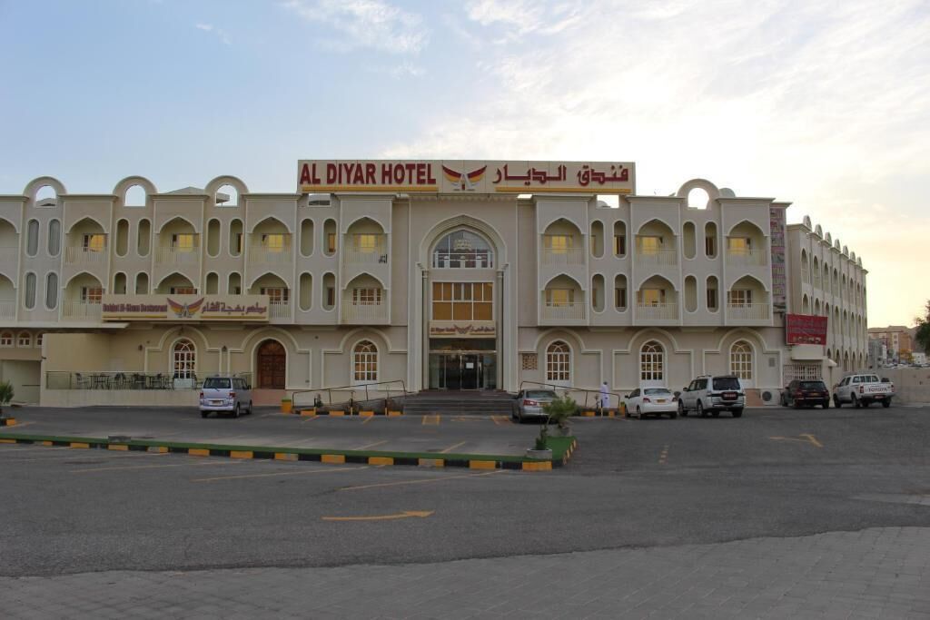 A hotel with a central entrance, multiple balconies, and a parking area.