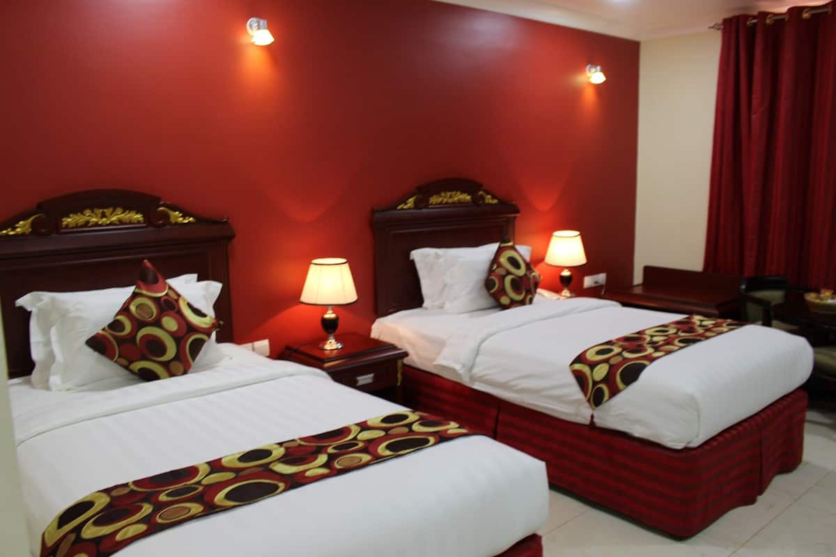 A hotel room with two beds, each with a red headboard, white bedding, and a patterned throw. There are bedside lamps and a red curtain on the window.