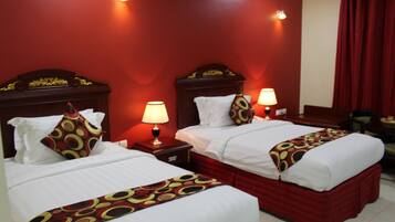 A hotel room with two beds, each with a red headboard, white bedding, and a patterned throw. There are bedside lamps and a red curtain on the window.
