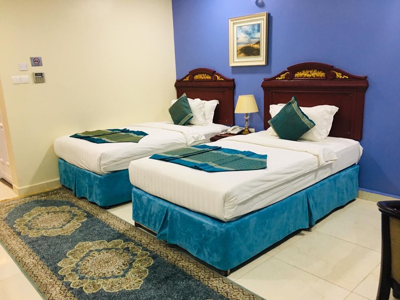 A hotel room with two beds, a blue accent wall, a painting, and a patterned rug.