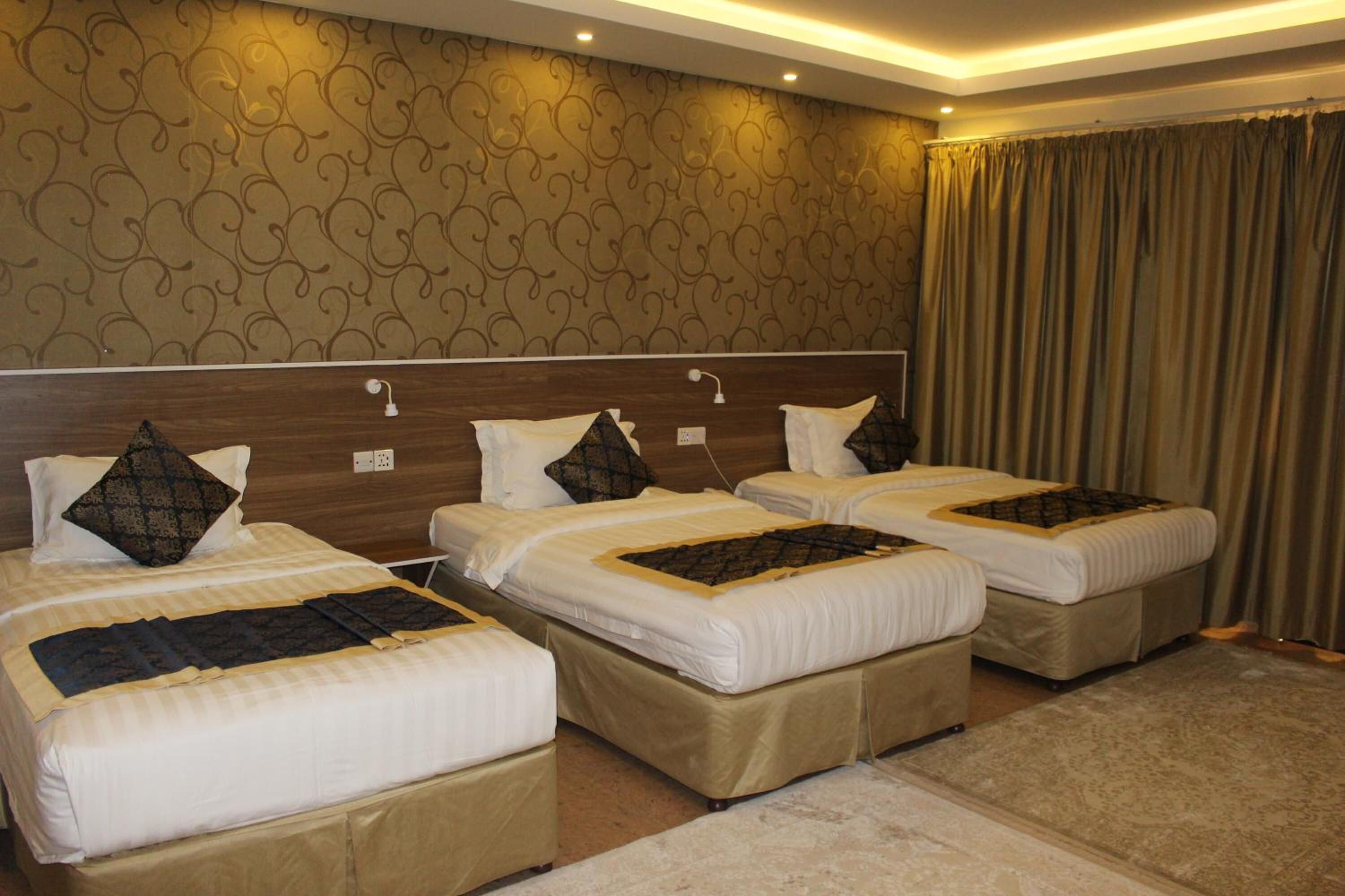 Une chambre d’hôtel avec trois lits simples, chacun doté d’une tête de lit, d’une table de chevet et d’un oreiller. La chambre est décorée d’un papier peint à motifs et possède une grande fenêtre avec des rideaux.