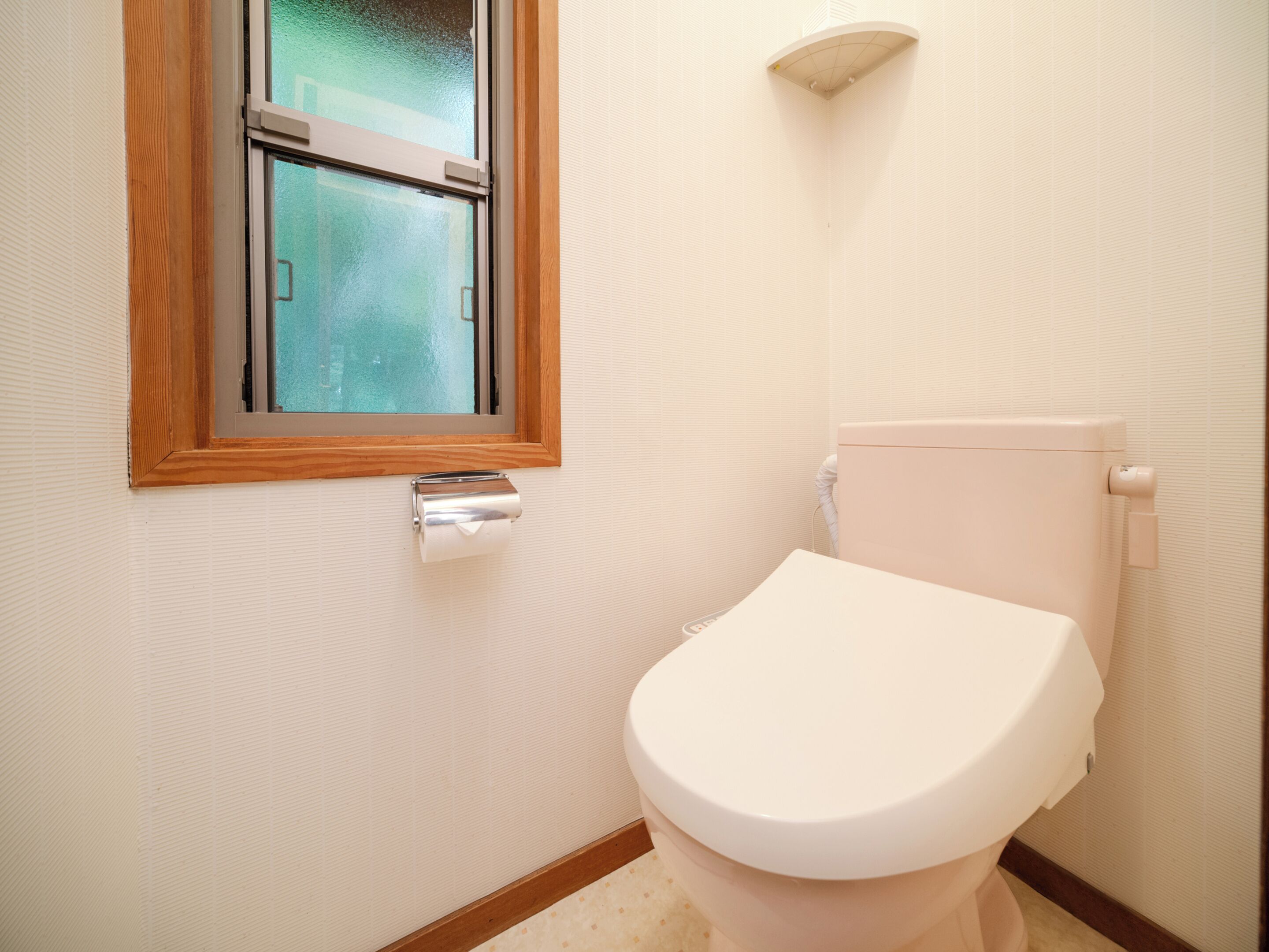 A toilet with a white lid, a roll of toilet paper, and a window with a wooden frame.