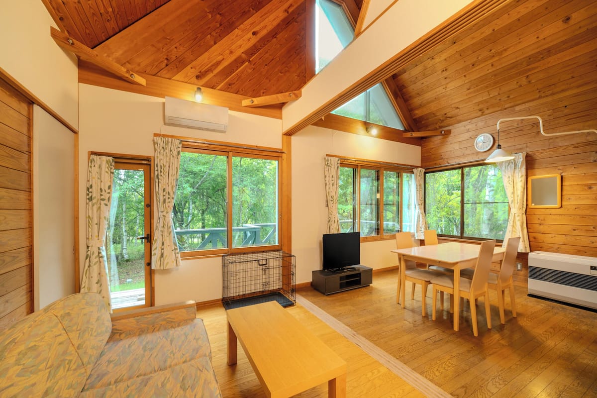 Interior de una cabaña con pisos de madera, zona de comedor, televisor y ventanas amplias.
