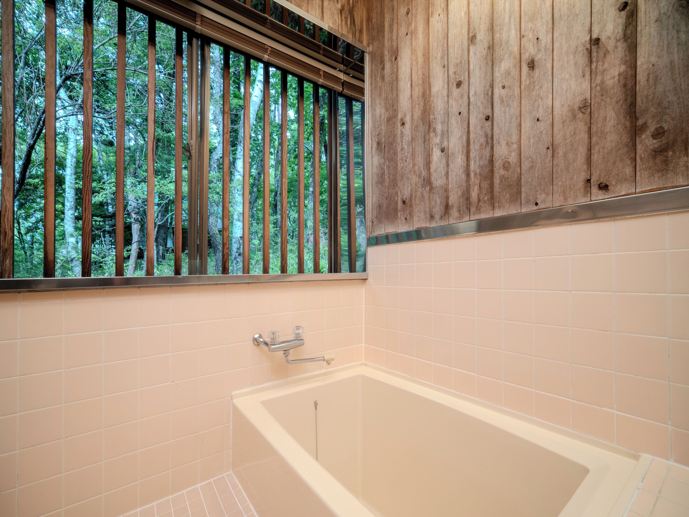 A bathroom with a bathtub, wooden window blinds, and a view of trees.