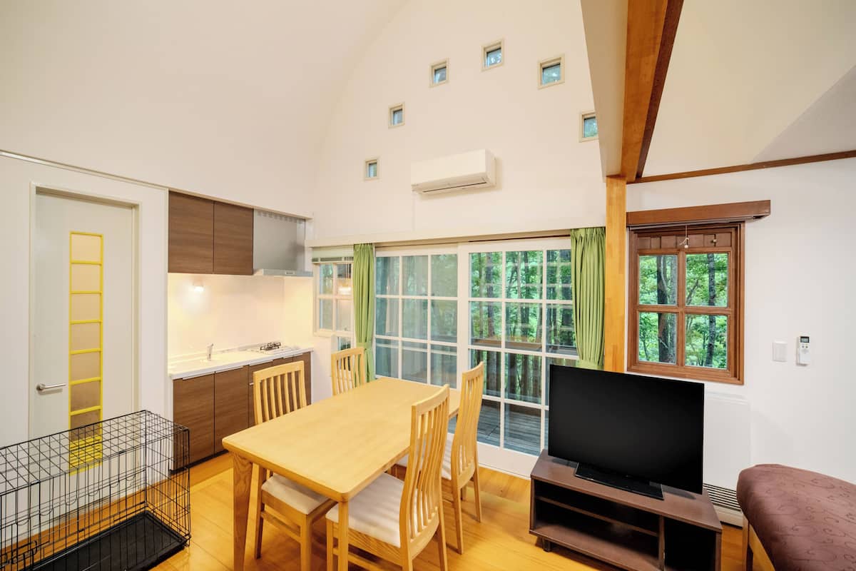 Un espacio habitable compacto con una mesa de comedor, sillas, un televisor, una zona de cocina y un ventanal con vistas a árboles.