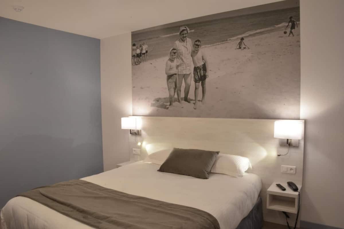 Habitación de hotel con cama, dos lámparas de noche, mesita de noche con teléfono y un cuadro colgado en la pared que muestra una playa.