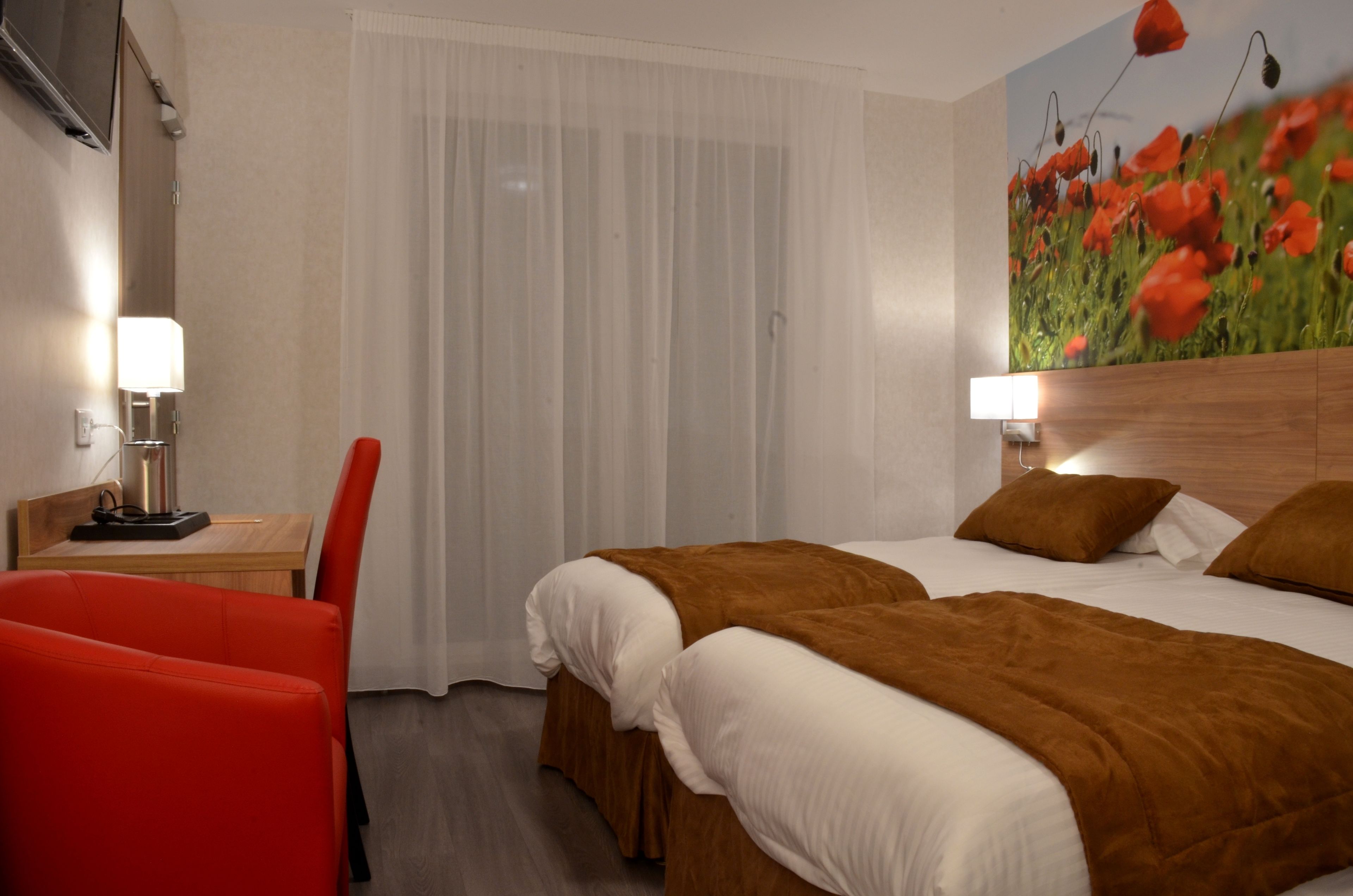 Une chambre d’hôtel avec un lit, deux tables de chevet avec des lampes, un fauteuil rouge et une fresque murale représentant des coquelicots.