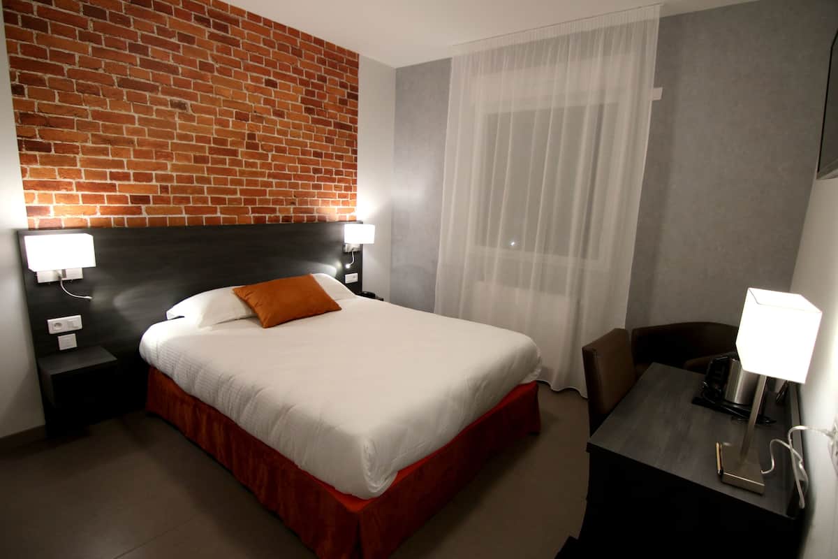 Habitación de hotel con pared de ladrillo, cama con ropa blanca y almohada naranja, escritorio con lámpara y una silla.
