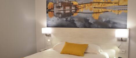 A hotel room with a bed, a headboard, bedside tables, and a painting of a bridge and trees on the wall.