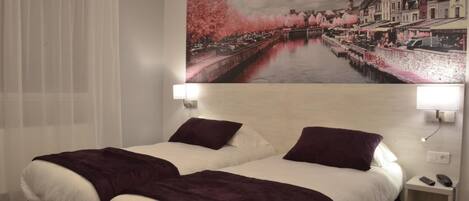 A hotel room with two beds, a large window with sheer curtains, a wall-mounted painting of a river scene, and bedside lamps.