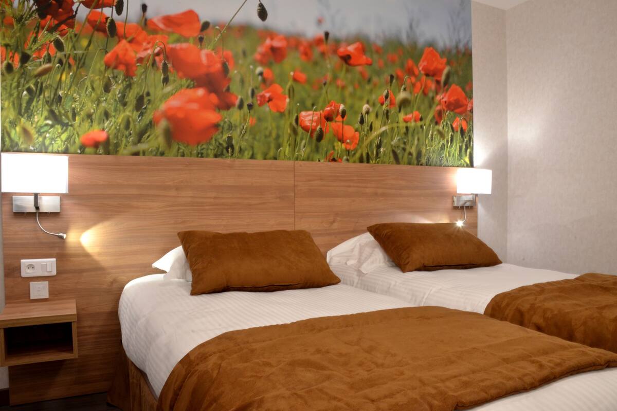 Habitación de hotel con un mural de un vasto campo de amapolas, cabecero de madera, dos camas con ropa de cama marrón y lámparas de noche.