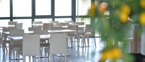 A spacious indoor area with multiple tables and chairs arranged in rows, large windows allowing natural light, and a potted plant in the foreground.
