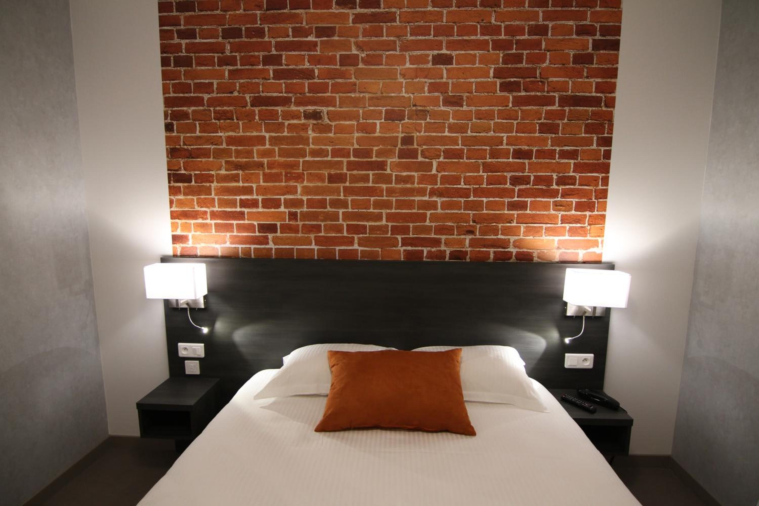Ein Hotelzimmer mit einer Wand im Backsteinmuster, einem schwarzen Kopfteil, einem weißen Bett mit einem braunen Kopfkissen und zwei Nachttischen mit Lampen.