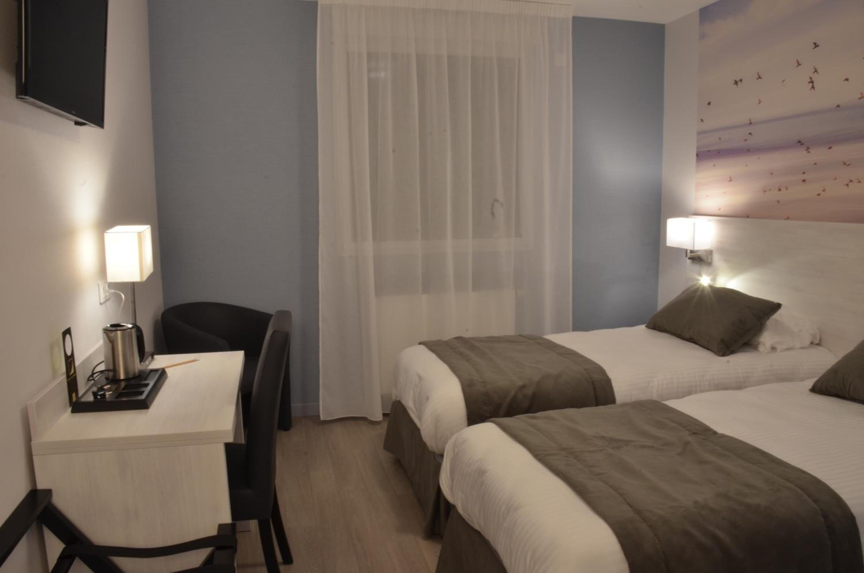 A hotel room with two beds, a desk with a lamp, a chair, and a TV mounted on the wall.