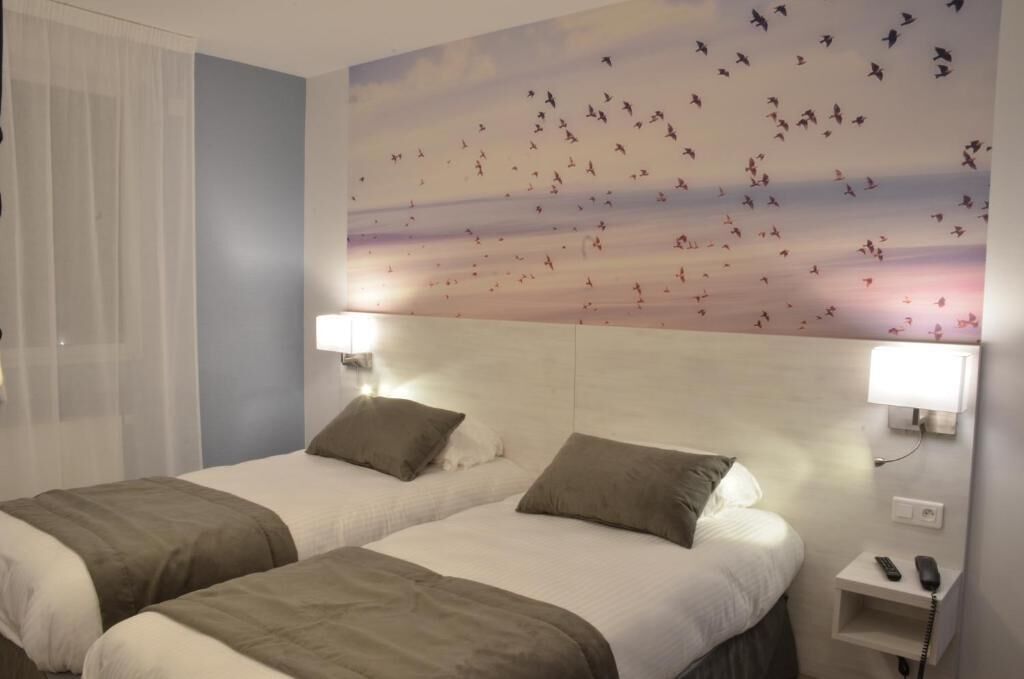 A hotel room with two beds, each with a pillow and a blanket. There is a wall-mounted lamp on the right side of the bed.