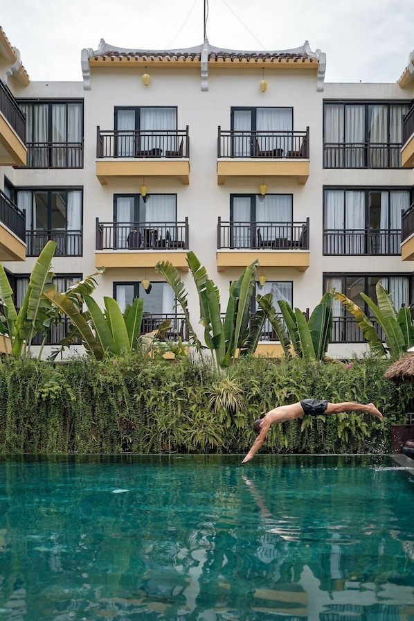 Une piscine où une personne est en train de plonger, entourée d’une végétation luxuriante et d’un bâtiment doté de balcons.