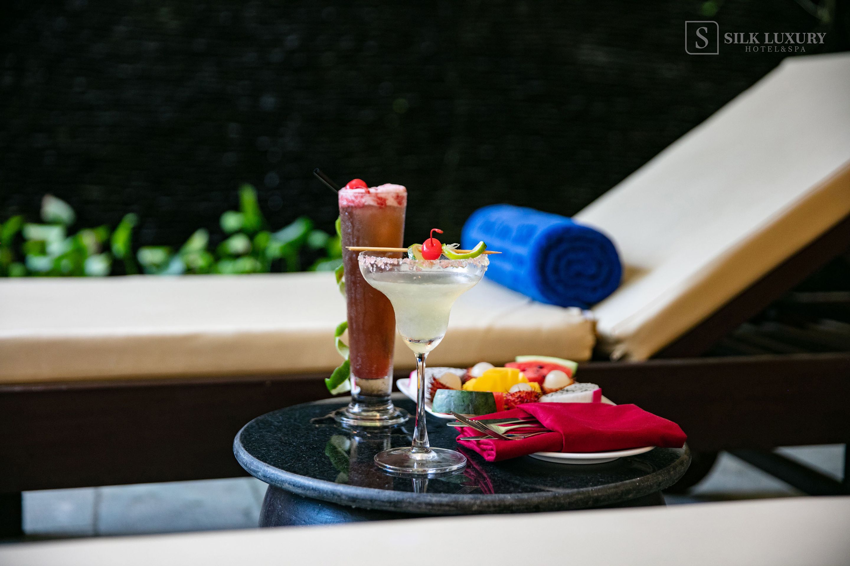 Un cocktail su un vassoio con guarnizione di frutta, un asciugamano arrotolato e un piatto di frutta su una poltrona prendisole.