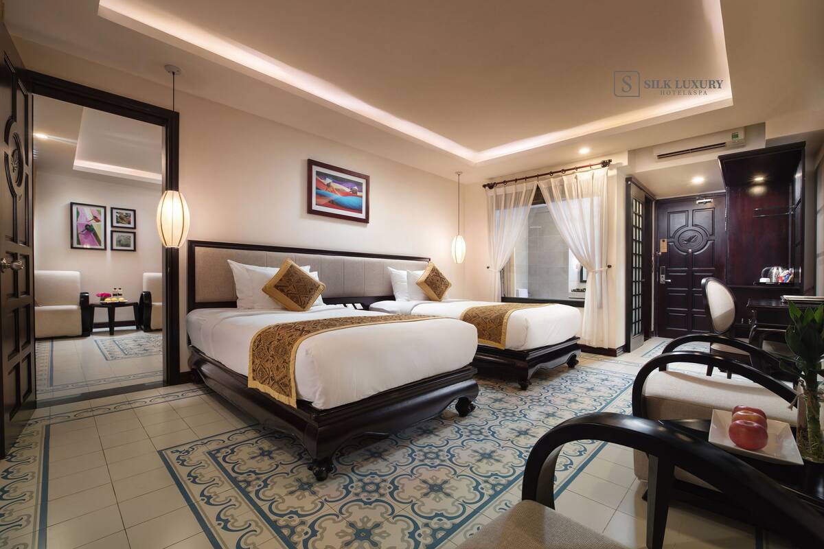 A hotel room with two beds, a sitting area with a sofa and table, a dining table with chairs, and a large mirror.