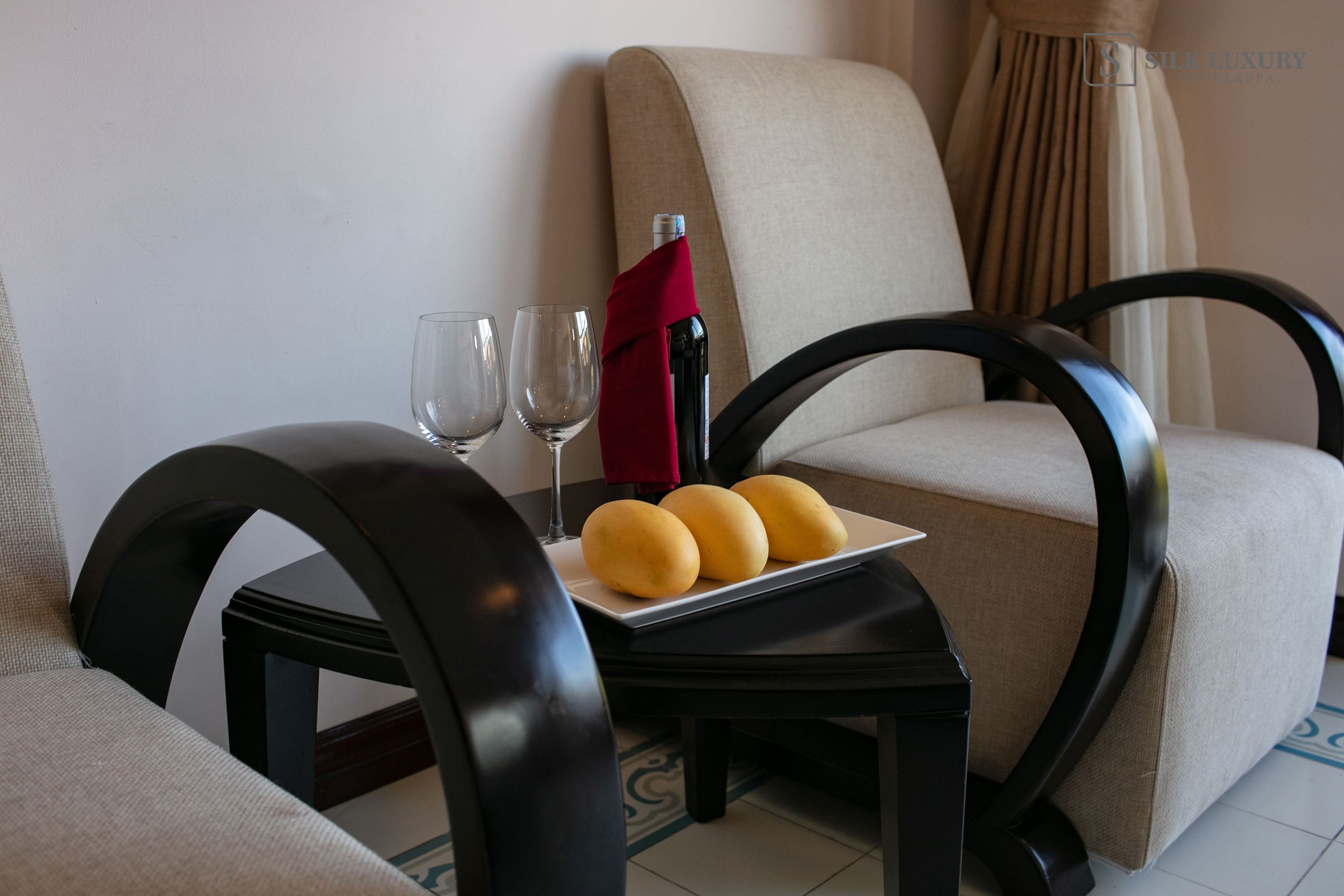 Una habitación de hotel con un sofá color beige, una mesita con una botella de vino, dos copas y un plato con tres pasteles.