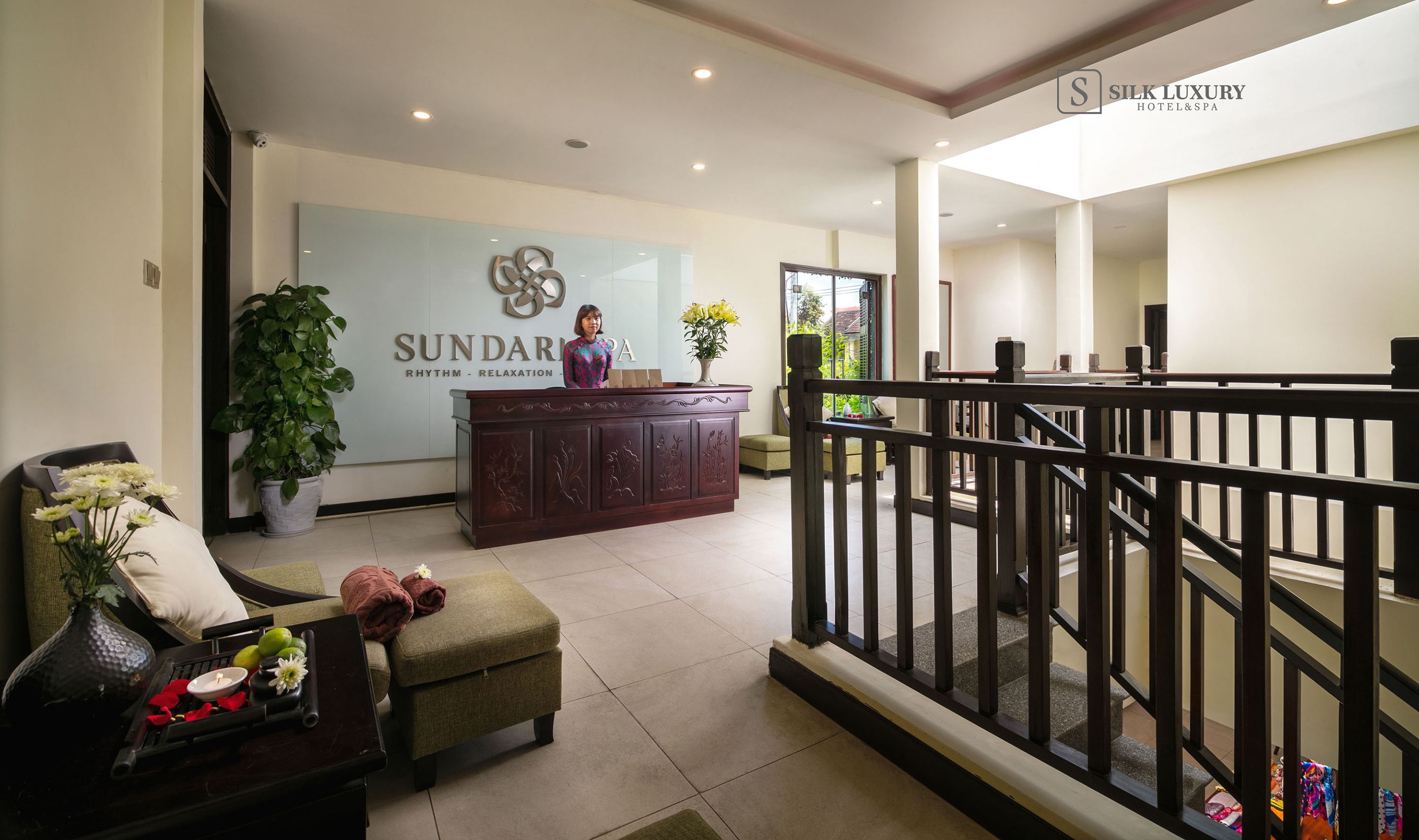 A reception area with a reception desk, a potted plant, a seating area with a sofa and a small table, and a staircase leading to an upper level.