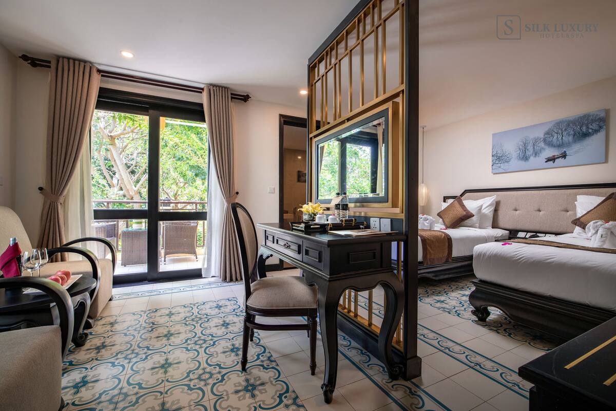 A hotel room with a desk, a chair, a bed, and a view of greenery through the window.