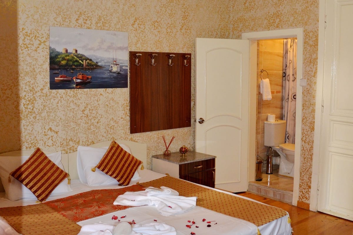Una cama doble con sábanas blancas y una colcha de motivos florales, techo de madera, un cuadro de barcos en la pared y una puerta de baño visible al fondo.