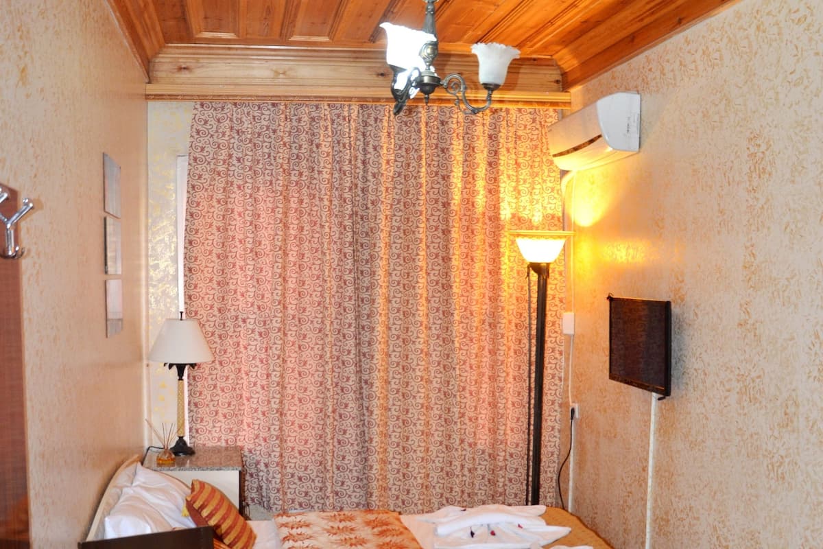 Una habitación pequeña y acogedora, con techo de madera, una cama con colcha estampada, una mesa con frutero y lámpara, y una alfombra roja con diseño.