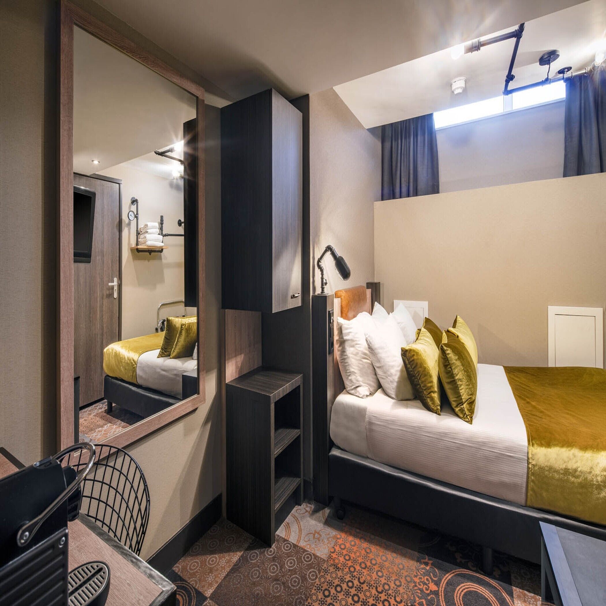 A modern hotel room with a bed, a desk, a chair, a mirror, and a window with curtains.