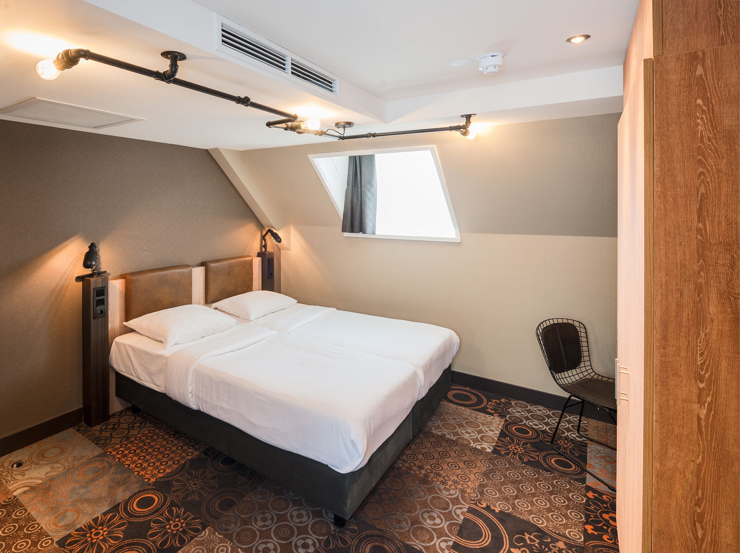 A modern hotel room with a neatly made bed, a chair, and a unique ceiling light fixture.
