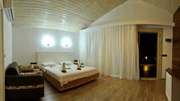 Un dormitorio con suelo de madera, una cama con ropa blanca, dos lámparas de pared y un gran ventanal con cortinas blancas.