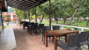 An outdoor patio with tables and chairs, a covered area, and a view of a garden and buildings. - Apella Hotel (Kemer)