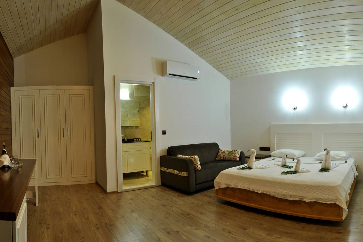 A bedroom with a wooden ceiling, a bed with decorative swans, a sofa, a wardrobe, and a bathroom visible through an open door.