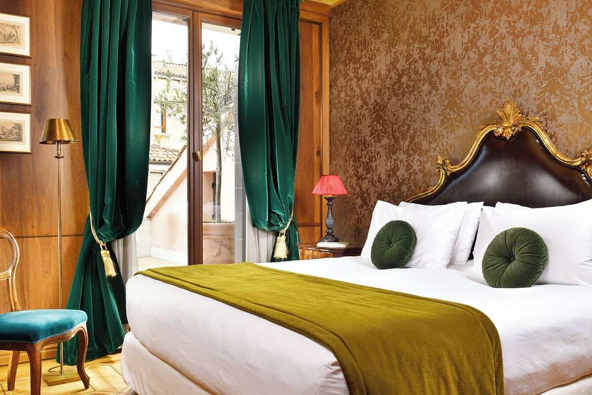 Habitación de hotel con una cama grande, cortinas verdes, una silla de madera y vistas al exterior.