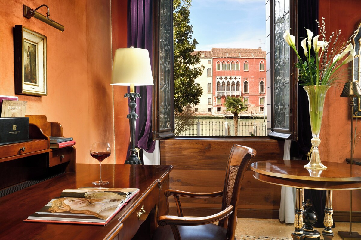 Una habitación con un escritorio de madera, una silla, una mesa, un jarrón con flores, una lámpara, un cuadro enmarcado y una ventana con vistas a un edificio.