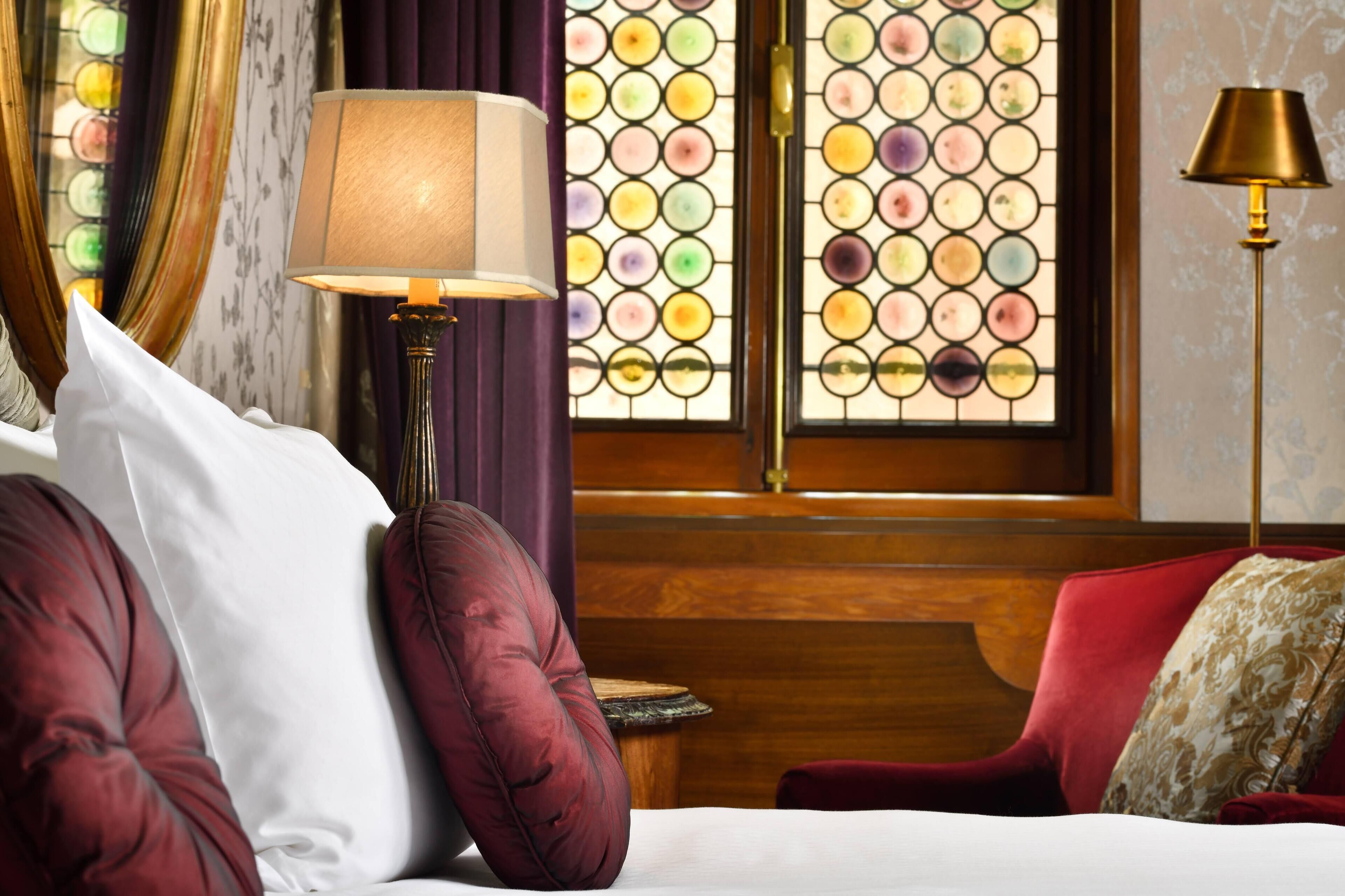 A hotel room with a bed, a red armchair, a wooden bench, and a stained glass window.