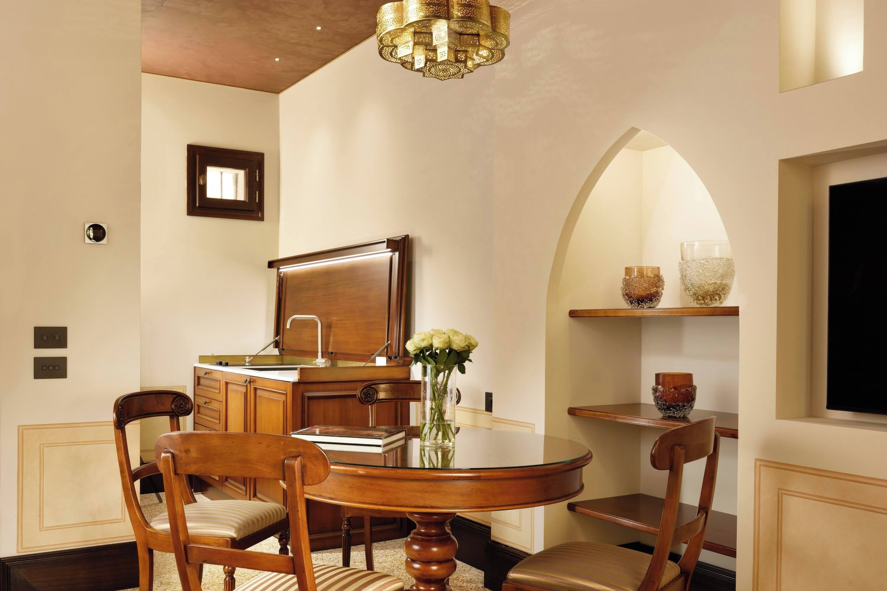 A dining area with a round wooden table, chairs, a vase with flowers, a chandelier, and a kitchen area with cabinets and a sink.