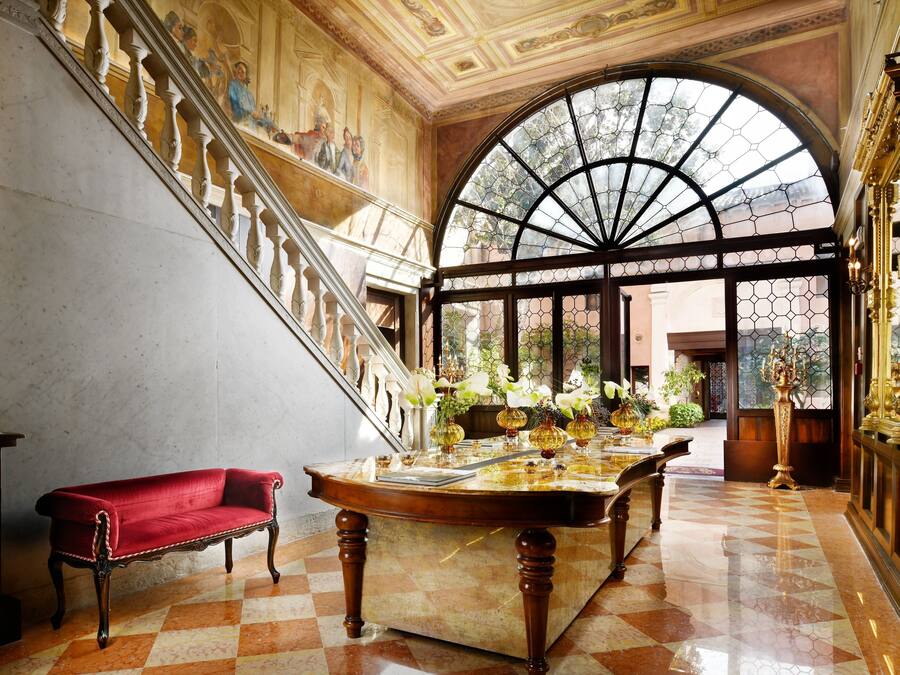 A grand staircase with a red sofa, a marble table with flowers, and a large arched window.