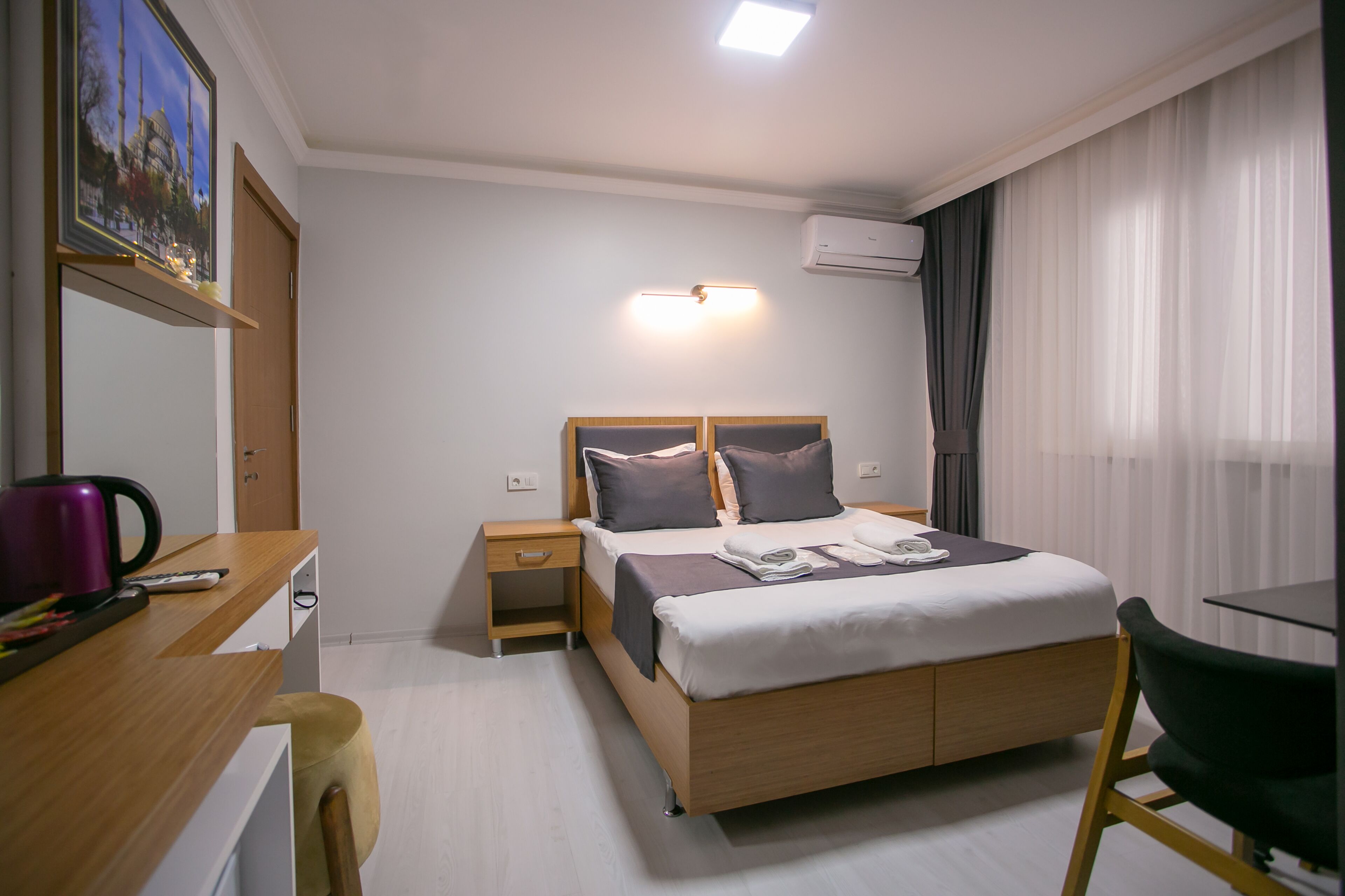 Ein Hotelzimmer mit einem hölzernen Bett, einem Schreibtisch mit Stuhl, einem Nachttisch, einer Wand-Klimaanlage und einem großen Fenster mit Vorhängen.