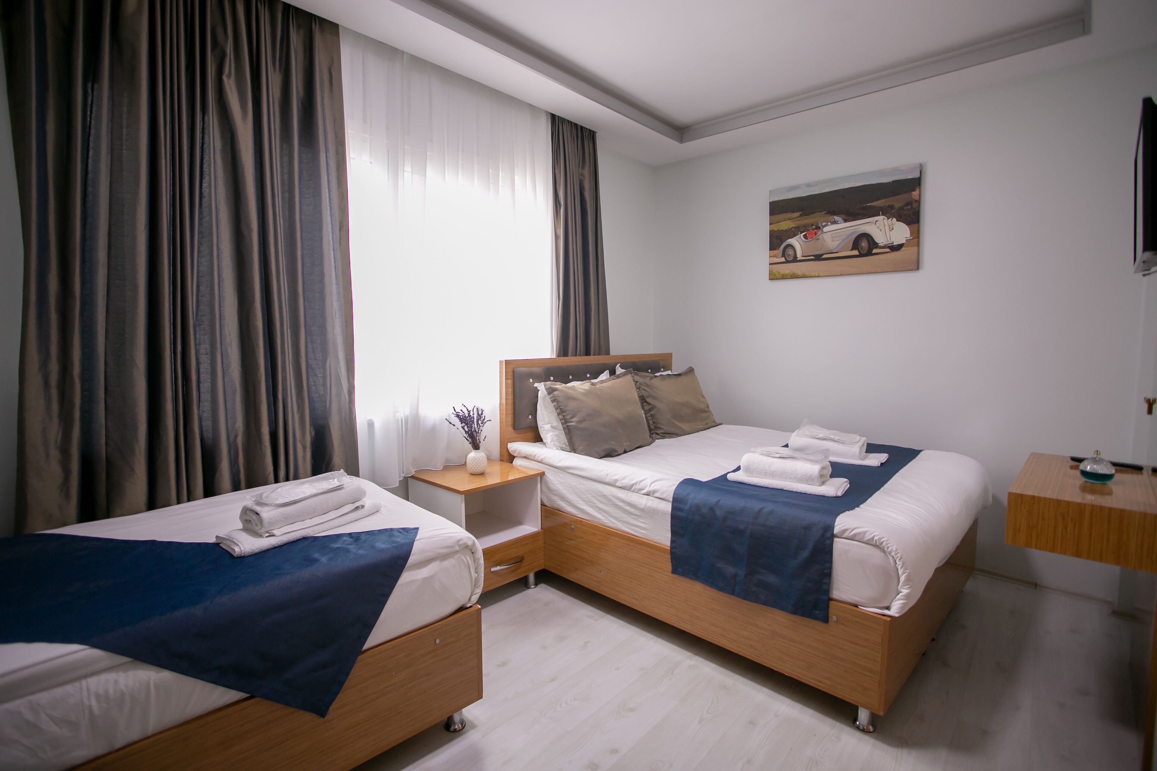 A hotel room with two beds, a nightstand, a bedside table, and a painting on the wall.