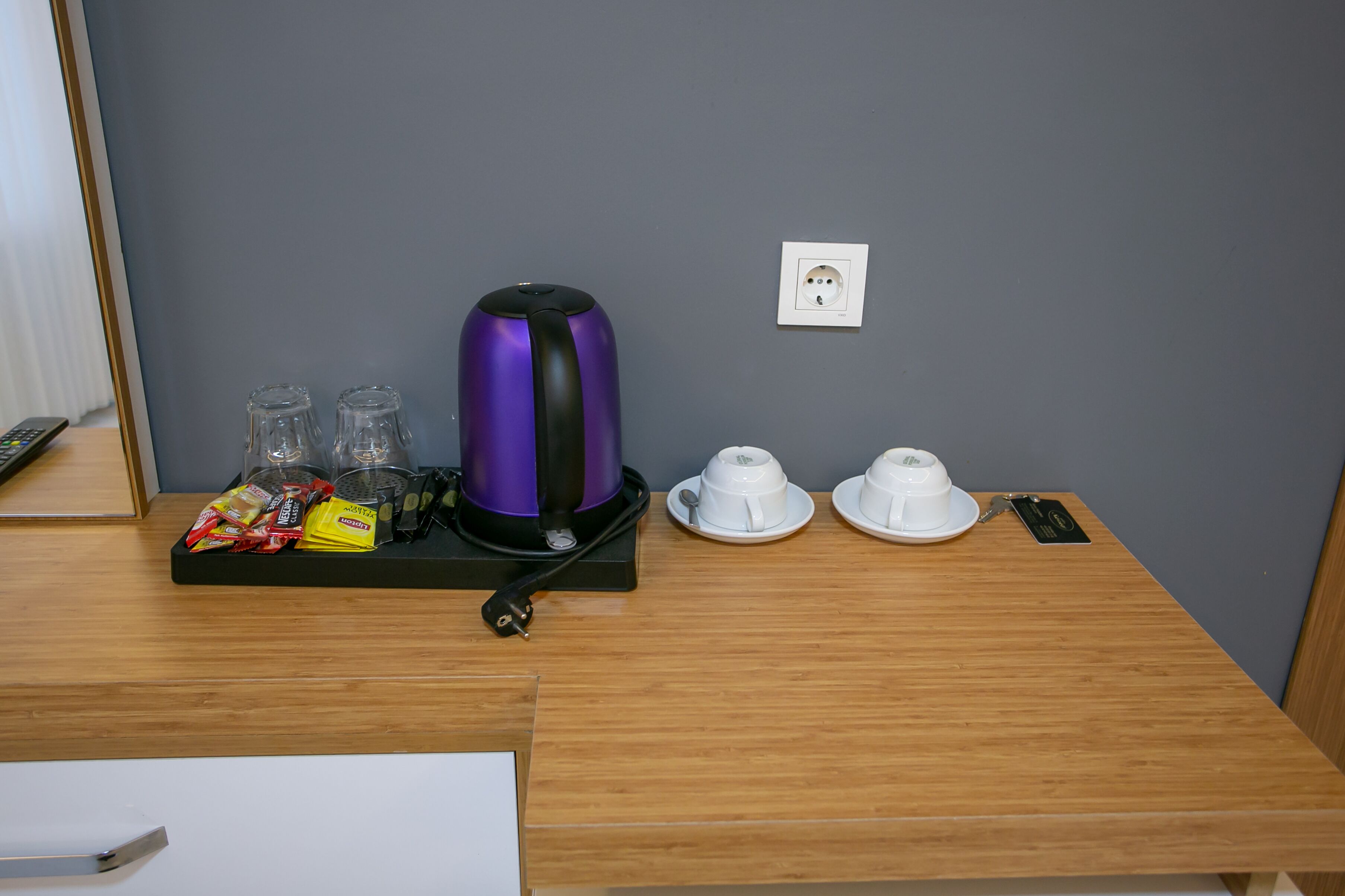 Une chambre d’hôtel avec une table en bois, une cafetière violette, deux tasses et un plateau contenant des en-cas et une télécommande.