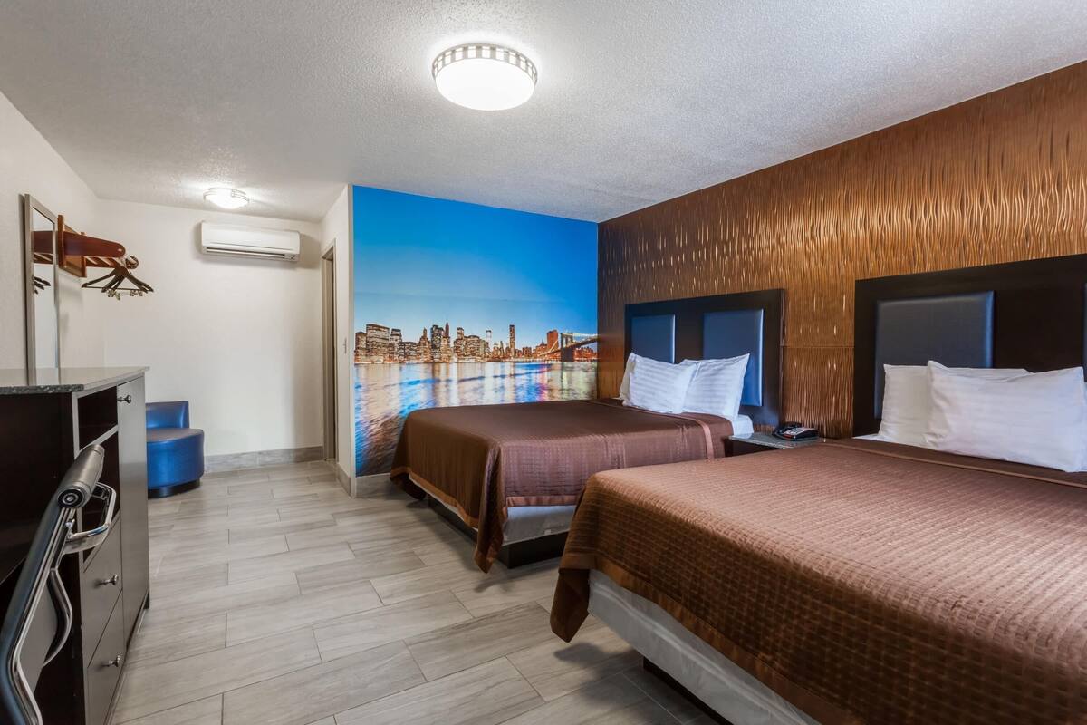 Habitación de hotel con dos camas, un mural de paisaje urbano, cabecera de madera y escritorio con espejo.