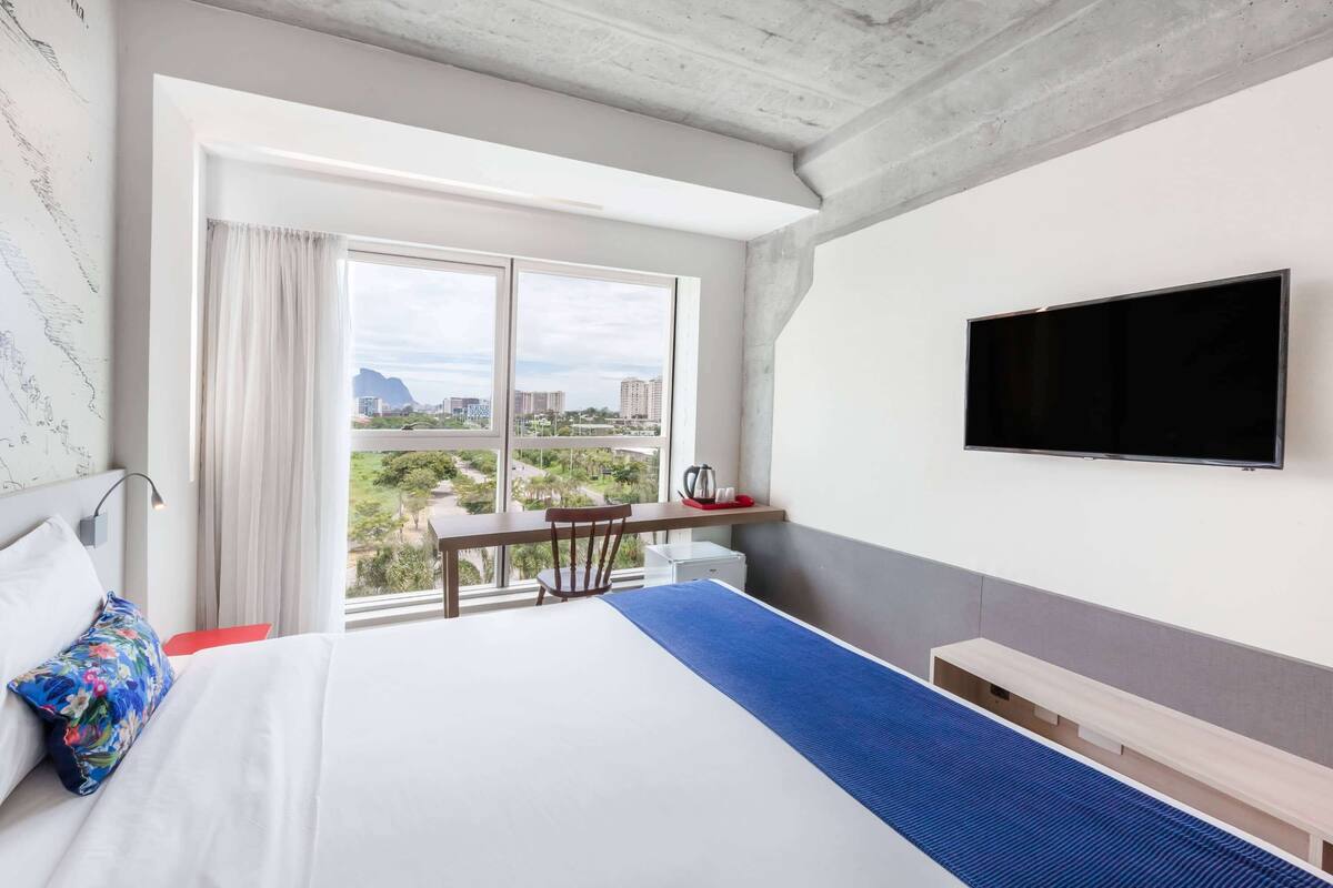 Ein Hotelzimmer mit einem großen Bett, einem Schreibtisch mit Stuhl, einem am Wand montierten Flachbildfernseher und einem Fenster mit Blick auf die Stadt.