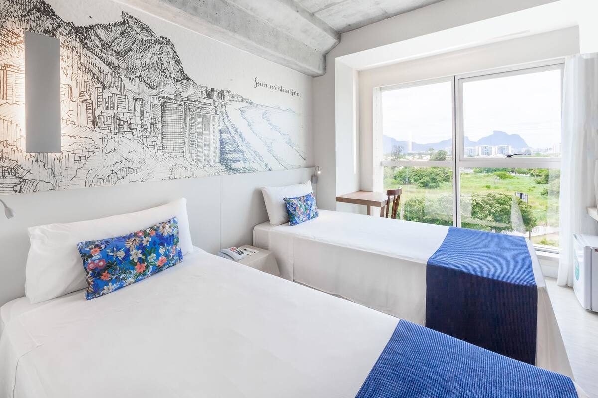 Ein Hotelzimmer mit zwei Betten, einem großen Fenster mit Blick auf die Stadt und einem Wandbild.